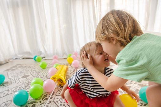 Adorable couple of laughing siblings playing on patterned floor covered with colorful balloons hugging and kissing