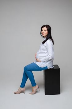 beautiful pregnant mum with black hair in a shirt and jeans sitting on a chair, picture isolated on grey background