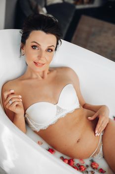 Portrait of gorgeous brunette woman expecting a baby posing in bath with roses in white lingerie, smiling at camera.