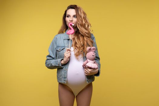 Funny young casual pregnant girl posing with hare toy and sweet candies isolated at yellow studio background. Beautiful fashion pregnancy woman enjoying motherhood medium long shot