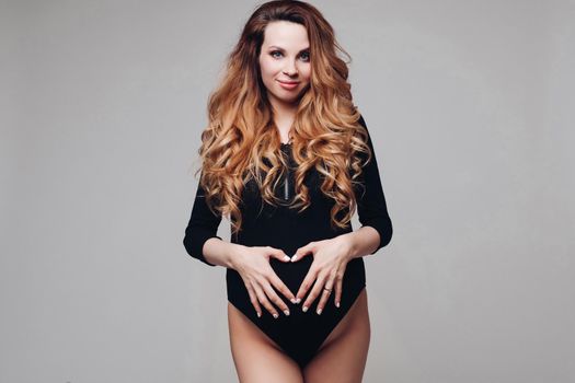 Waist up of happy young beautiful pregnant woman holding palms on her stomach isolated on grey background