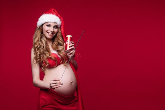 11 27 2019 Belarus Minsk: Adorable pregnant young woman in Santa Claus suit posing holding big tummy isolated at red studio background. Smiling festive future mother enjoying motherhood having positive emotion medium long shot