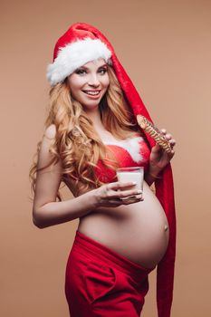 Smiling beautiful pregnant lady in Santa Claus hat soaking gingerbread in milk on beige background. Christmas and New Year concept