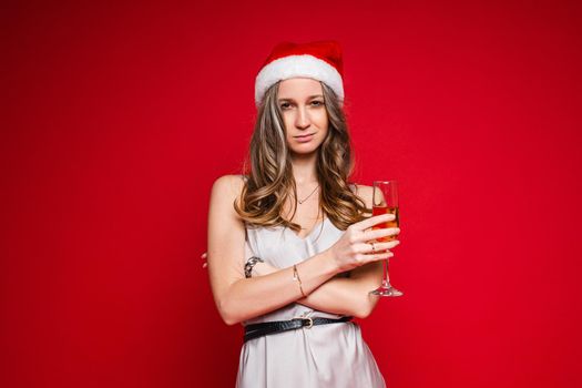 Pretty young female in santa hat and festive dress posing holding champagne glass on red background, copy space. High quality photo