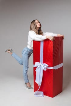 Full length portrait of attractive young woman with fair hair in casual white sweater, jeans and heels hugging big red wrapped gift with white bow with pouting lips.