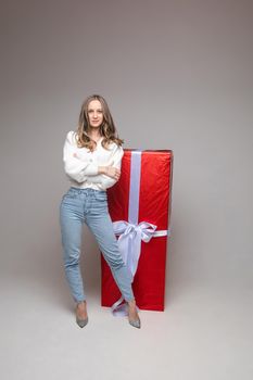 Happy beautiful lady standing in front of the big red gift box, isolated on grey background. Saint Valentine Day concept