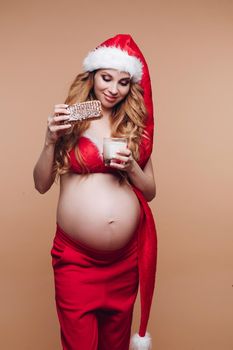 Smiling pregnant girl in Santa Claus suit eating gingerbread cookies with milk having fun full shot. Beautiful festive future mother celebrating Christmas holiday isolated at nude studio background