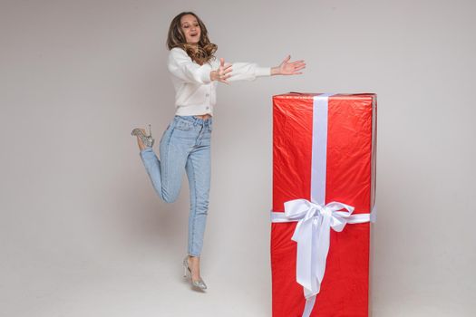 Full length stock photo of cheerful jumping girl in heels with outstretched arms with giant gift in red paper with white bow on white background.