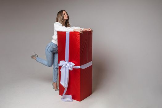 Full length portrait of attractive young woman with fair hair in casual white sweater, jeans and heels hugging big red wrapped gift with white bow with pouting lips.