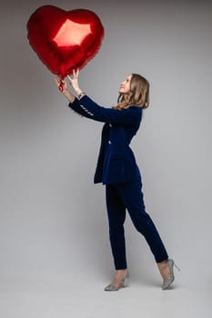 Full length of happy lady in suit holding heart shaped balloon and looking at it, isolated on grey background. Saint Valentine Day concept