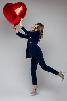 Full length of happy lady in suit holding heart shaped balloon and looking at it, isolated on grey background. Saint Valentine Day concept