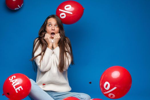 Portrait of shocked brunette woman in white sweater pulling her collar up to opened mouth and looking up. Red air balloons with sale and discount sign flying around.