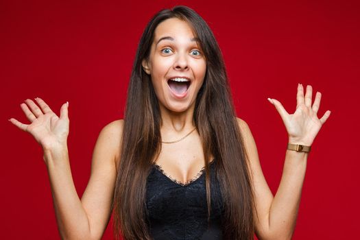 Portrait of astonished and surprised beautiful brunette woman in black top looking at camera with wide eyes and bent raised arms on red background. Surprised with cool present.