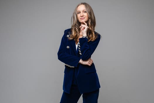 Portrait of beautiful adult woman in stylish trouser suit holding hand near her chin while looking at camera, isolated on grey background