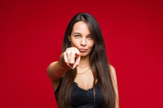 Waist up of smiling pretty woman posing in studio and pointing her finger at camera, isolated on red background. Party concept. Copy space