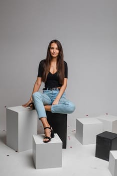 beautiufl caucasian woman with charming appearance sits on a white cube and looks to te camera, picture isolated on white background