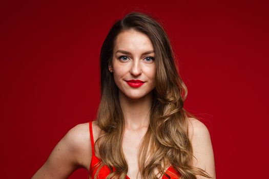 Studio portrait of stunning young adult woman with long wavy brown hair smiling at camera with plump red lips. Isolated on red.