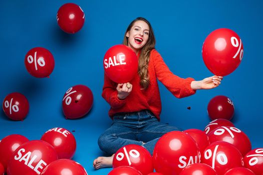 Joyful young girl sitting on the floor on blue studio background with red balloons, sale and discount concept. High quality photo