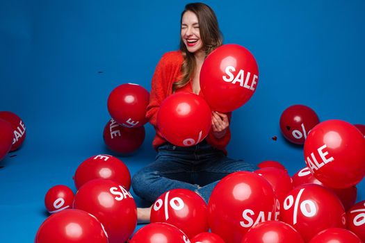 Smiling pretty lady holding two red balloons in studio, isolated on blue background. Shopping holiday concept