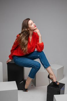 Studio shot of attractive brunette with red lips in stylish casual red pullover and denim jeans and black heels sitting on black and white cubes smiling at camera.