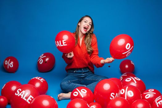 Portrait of cheerful brunette in red blouse and jeans sitting with crossed legs and holding red balloon with discount sign and sale word.