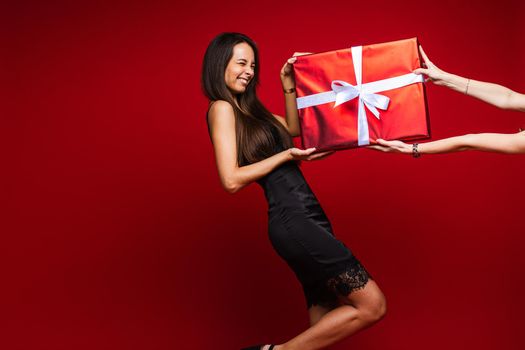 Portrait of laughing brunette in black dress with lace rim getting giant present in red paper and white bow on Valentine s day.