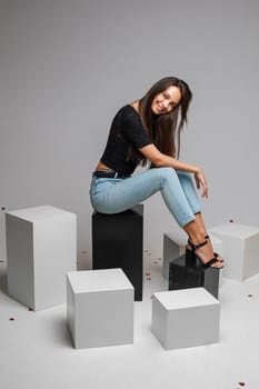 Smiling pretty young woman posing in studio while sitting on black boxes, isolated on grey background. Copy space