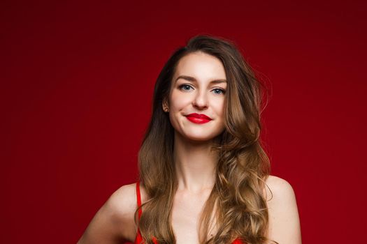 Studio portrait of stunning young adult woman with long wavy brown hair smiling at camera with plump red lips. Isolated on red.