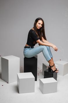 Smiling young fit girl with long hairs posing sitting on black and white cubes on gray studio background with copy space. High quality photo