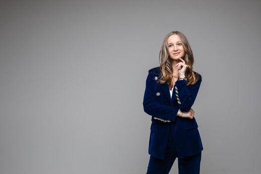 Studio portrait of pretty smiling fair-haired Caucasian model in trendy dark blue suede jacket and trousers with bent arm at chin. Copy space.