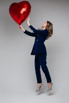 Full length of happy lady in suit holding heart shaped balloon and looking at it, isolated on grey background. Saint Valentine Day concept