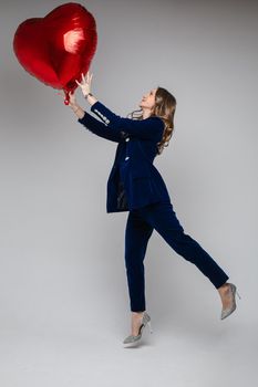 Full length of attractive Caucasian woman in blue velvet suit with jacket and trousers and high sparkling heels holding red heart-shaped symbol of love balloon. Isolated.