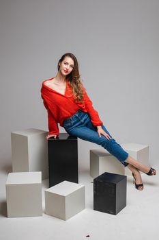 Studio shot of attractive brunette with red lips in stylish casual red pullover and denim jeans and black heels sitting on black and white cubes smiling at camera.