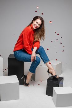 Joyful smiling young girl under festive confetti wearing red sweater, jeans posing on black and white cubes in studio. High quality photo