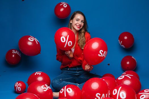 Smiling pretty lady holding two red balloons in studio, isolated on blue background. Shopping holiday concept