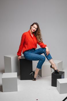 Studio shot of attractive brunette with red lips in stylish casual red pullover and denim jeans and black heels sitting on black and white cubes smiling at camera.