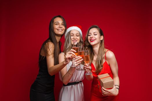 Happy young women friends with gift and champagne celebrating holiday together on party on red background, copy space. High quality photo