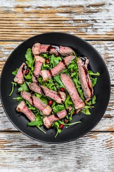 Sliced grilled beef steak with arugula leaves salad. White wooden background. Top view.