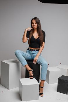 Full length of beautiful smiling brunette in black top and blue jeans and heels sitting on black cubes and looking at camera with bent head.