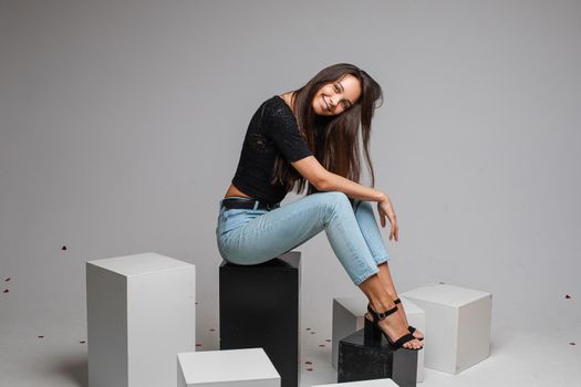 Full length of beautiful smiling brunette in black top and blue jeans and heels sitting on black cubes and looking at camera with bent head.