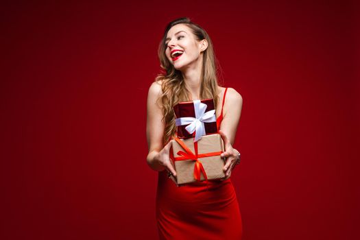 Portrait of attractive young lady in red silk dress holding two small wrapped presents in arms and smiling away. Isolated on red background.