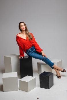 Studio shot of attractive brunette with red lips in stylish casual red pullover and denim jeans and black heels sitting on black and white cubes smiling at camera.