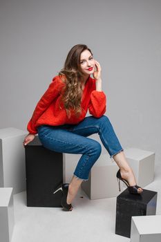 Studio shot of attractive brunette with red lips in stylish casual red pullover and denim jeans and black heels sitting on black and white cubes smiling at camera.