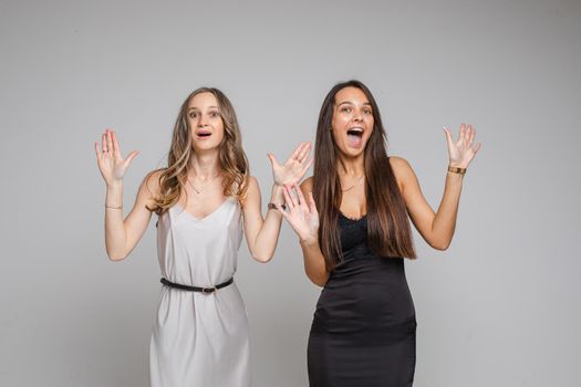 Two pretty women standing in studio pointing their fingers up, isolated on grey background. Party concept. Copy space