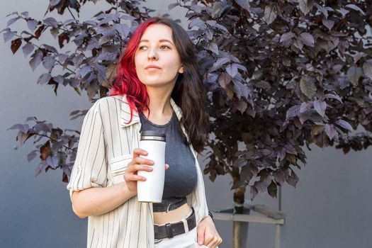Young woman with creative colored black and red hair holding a white reusable insulated mug for coffee to go in her hands.