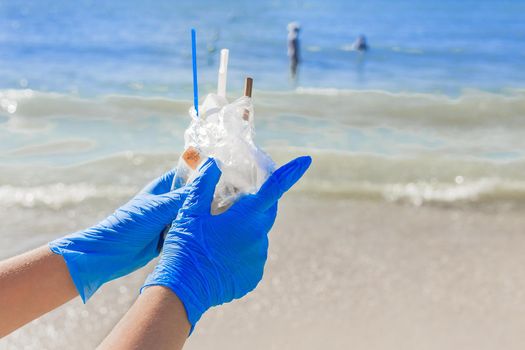 Hands in blue protective household gloves hold a pile of garbage in a bag against the sea. Waste disposal.