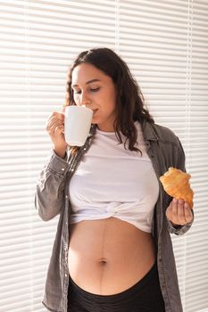 Healthy beautiful pregnant woman drinking tea and eating croissant during lunch. High-calorie nutrition while waiting for birth of baby