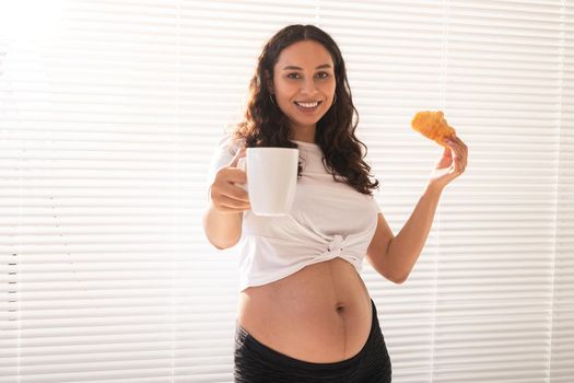 Beautiful pregnant woman holding croissant and cup of coffee in her hands during morning breakfast. Good health and positive attitude while expecting baby