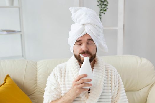 Bearded male with towel on his head applying body lotion cream. Guy smile when smell lotion cream on hand. Spa, body and skin care for man concept.