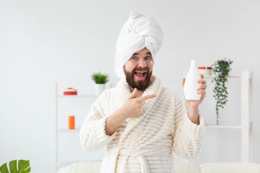 Beauty portrait of handsome beautiful guy in towel and bathrobe holding tube of body cream lotion in hand. Spa, body and skin care for man concept.